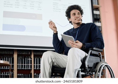 Low angle view of self-confident Middle Eastern student with disability holding digital tablet doing presentation at event in college library - Powered by Shutterstock