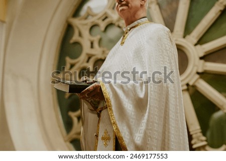 Low angle view of senior Catholic priest wearing vestment holding Bible book standing against round window at church balcony