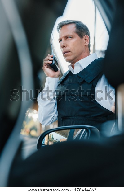 low angle\
view of serious mature male police officer in bulletproof vest\
talking on radio set near car at\
street