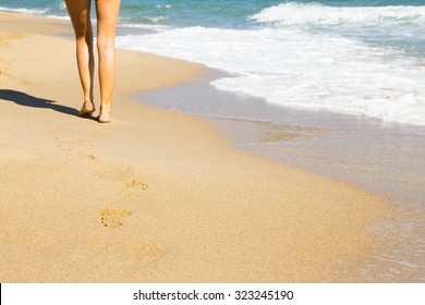 Low Angle View Of The Sexy Bare Legs Of A Woman Walking Away Along A Beach Across The Golden Sand At The Edge Of The Water With Gentle Surf, Copy Space In The Foreground