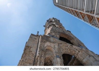 A low angle view shows the damaged Kaiser Wilhelm Memorial Church tower, brick arches and gold clock. Scaffolding and blue sky emphasize restoration and historic urban texture.
