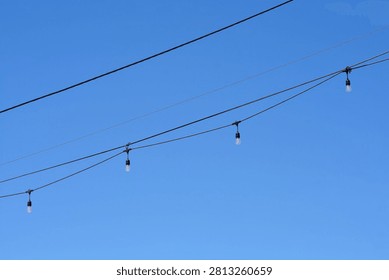 A low angle view shows strings of light bulbs hanging from outdoor overhead wires against a clear bright blue sky
