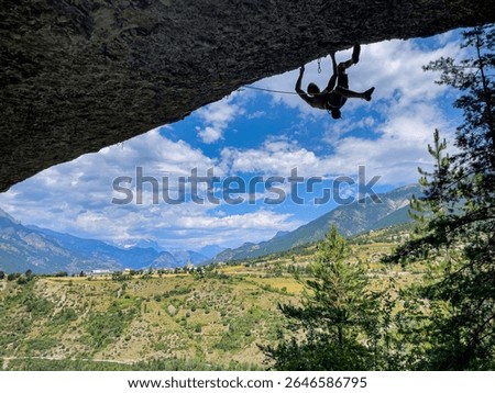 LOW ANGLE VIEW, SILHOUETTE: Athletic climber hangs from rocky overhang, with view of green valley and majestic mountains under cloudy blue sky. Dramatic scale and beauty of outdoor climbing experience
