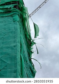 Low angle view of a skyscraper under construction wrapped in torn green safety netting against a cloudy sky