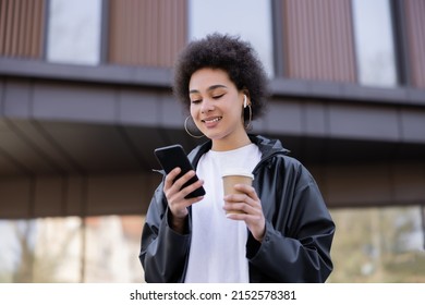 Low Angle View Of Smiling African American Woman In Earphone Holding Paper Cup And Smartphone Outside