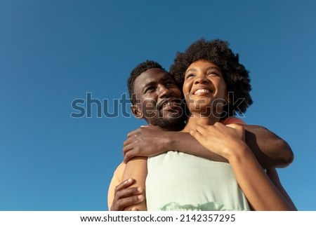 Low angle view of smiling african american man hugging girlfriend from behind against blue sky. unaltered, copy space, lifestyle, love, togetherness and holiday concept.