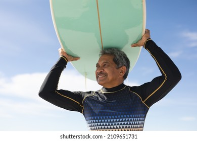 Low Angle View Of Smiling Biracial Senior Man Carrying Surfboard On Head Against Sky At Sunny Beach. Water Sport And Active Lifestyle.