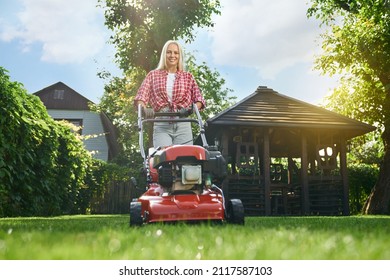 Low angle view of smiling caucasian woman with blond hair using electric lawn mower while working at garden. Attractive female in casual clothes cutting grass with modern gardening equipment.