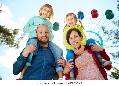 Low Angle View Of Smiling Family In Amusement Park, Mother And Father Carrying Their Two Adorable Blond Girls On Shoulders And Looking At Camera During Warm Autumn Weekend, Ferris Wheel In Background
