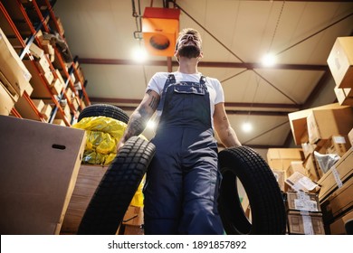 Low Angle View Of Smiling Hardworking Blue Collar Worker In Overalls Carrying Tires And Walking In Storage In Import And Export Firm.