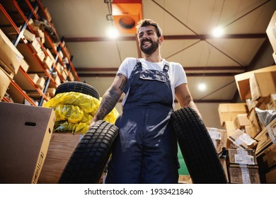 Low Angle View Of Smiling Hardworking Blue Collar Worker In Overalls Carrying Tires And Walking In Storage In Import And Export Firm.