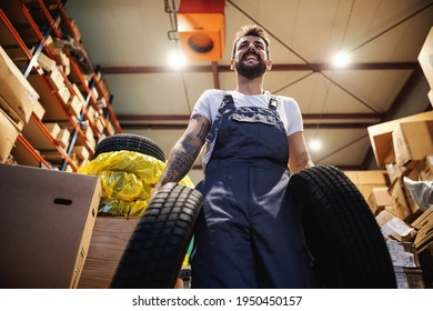 Low Angle View Of Smiling Hardworking Blue Collar Worker In Overalls Carrying Tires And Walking In Storage In Import And Export Firm.