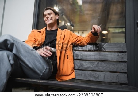 Low angle view of smiling and stylish young man holding cigarette and paper cup on bench near cafe