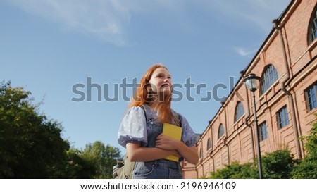 Low angle view of smiling teenage girl with book and backpack standing outside school building, looking around and smiling. Education, back to school concept