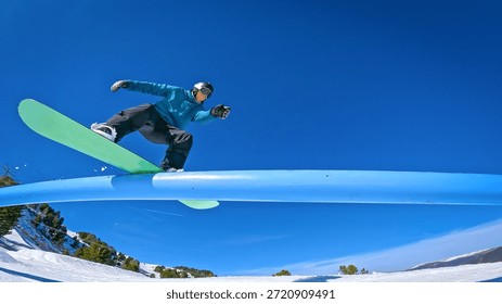 LOW ANGLE VIEW: Snowboarder doing a stylish boardslide trick on a blue rail. Freestyle rider enjoys doing laps through a fun snowpark at sunny ski resort during winter holidays in snowy Austrian Alps.