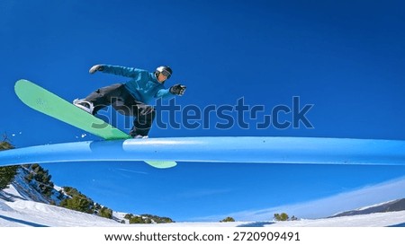 LOW ANGLE VIEW: Snowboarder doing a stylish boardslide trick on a blue rail. Freestyle rider enjoys doing laps through a fun snowpark at sunny ski resort during winter holidays in snowy Austrian Alps.