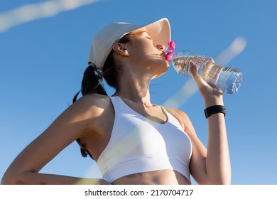 low angle view of sportive woman in baseball cap and sports bra drinking water against blue sky