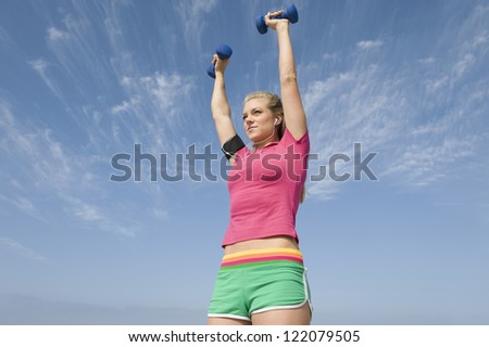 Low angle view of sporty young woman exercising with hand weights while listening music against sky