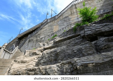 Low angle view of a stone retaining wall and rugged rock cliff with stairs, railings and a small green tree under a bright blue sky.