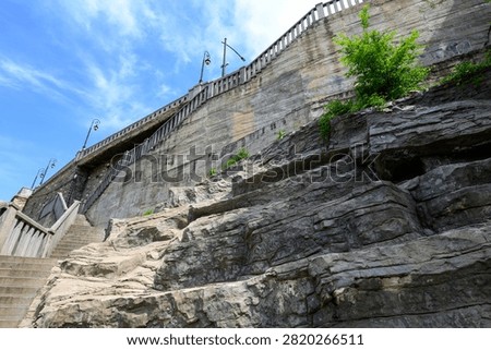Low angle view of a stone retaining wall and rugged rock cliff with stairs, railings and a small green tree under a bright blue sky.