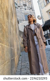 Low Angle View Of Stylish African American Woman In Trench Coat Walking Near Ancient Wall In Prague