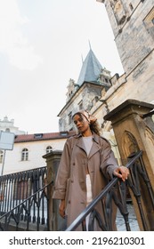 Low Angle View Of Stylish African American Woman In Trench Coat Near Old Town Hall Tower In Prague