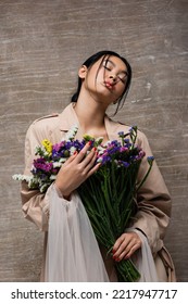 Low Angle View Of Stylish Asian Woman In Trench Coat Holding Wildflowers On Abstract Brown Background 