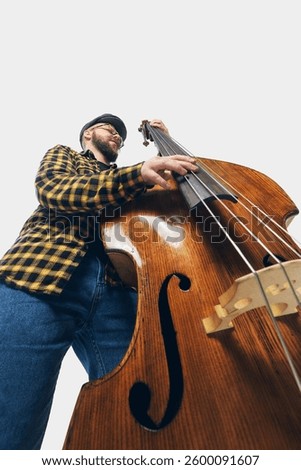 Low angle view of stylish double bass player in checkered shirt leaning into deep groove, fingers playing over rich wooden strings. Concept of music, jazz, concert, festival, rhythm, instruments.