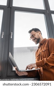 low angle view of successful and bearded businessman in eyeglasses and stylish shirt standing near window in modern office and smiling while using laptop, corporate lifestyle