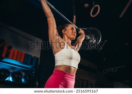 Low angle view of a sweaty fit young female in sportswear exercising in a dark modern gym while lifting heavy weights. Workout sessions. Woman having a fitness training indoor. Copy space.