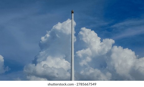 Low angle view of a tall empty white flagpole with a gold finial against a blue sky with fluffy white cumulus clouds