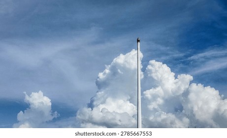 Low angle view of a tall empty white flagpole with a gold finial against a blue sky with fluffy white cumulus clouds