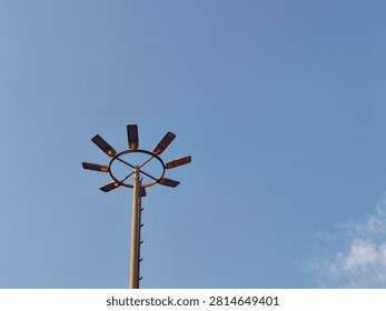 Low angle view of a tall high mast lighting pole with multiple floodlights arranged in a circular ring pattern against a clear blue sky.