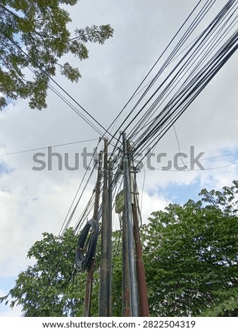 Low angle view of tangled electrical wires and fiber optic cables on poles with green trees background.

