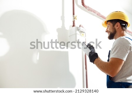 Low angle view of technician, heating fitter in hard hat and protective gloves making notes while checking gas consumption at gas pipe and meter counter
