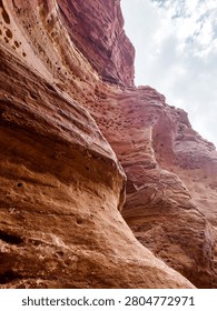 Low angle view of towering red sandstone cliffs under a cloudy sky in a desert canyon.