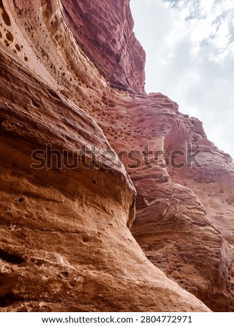 Low angle view of towering red sandstone cliffs under a cloudy sky in a desert canyon.
