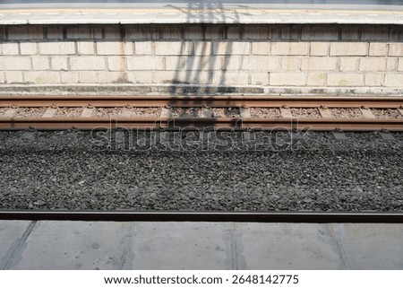 Low angle view of train tracks, ballast, and a brick wall with shadows cast by overhead structures.