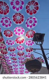 A low angle view of translucent umbrellas with pink petal motifs hanging against a deep blue sky. Sunlight filters through the canopy near an old fashioned iron street lamp and a flower basket.