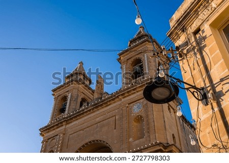 Low angle view of twin bell towers and facade in Marsaxlokk, Malta, lit by festive bulbs and late sun. Warm limestone, Baroque details, wires and a streetlamp suggest an event.