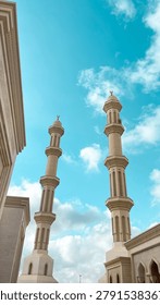 Low Angle View of Twin Mosque Minarets Against a Blue Cloudy Sky. A dramatic low-angle architectural photograph focusing on two towering, detailed minarets of a modern white mosque. Reaching up toward a vibrant turquoise blue sky filled with bright white clouds, this striking vertical composition is perfect for Islamic religious design, travel brochures, cultural heritage projects, and Ramadan or Eid holiday graphics.