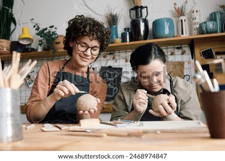 Low angle view of two diverse women sitting at table in pottery workshop making ceramic bowls using clay carving tools