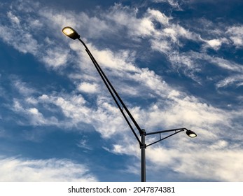 Low Angle View Of Two Suburban Street Lights, Each On A Long Arm Attached To The Same Pole, Against An Evening Sky With Cirrus Clouds