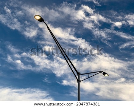 Low angle view of two suburban street lights, each on a long arm attached to the same pole, against an evening sky with cirrus clouds