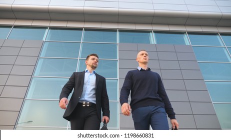 Low angle view of two young successful colleagues in formal wear walking with briefcases from office building after work. Handsome businessmen going with bags to home after job in city. Close up.