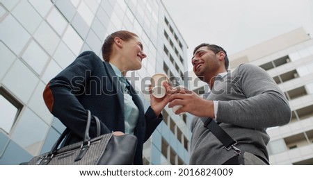 Low angle view of two young intercultural colleagues with coffee interacting outdoors. Caucasian businesswoman and indian businessman in smart casual wear drink coffee-to-go and discuss downtown