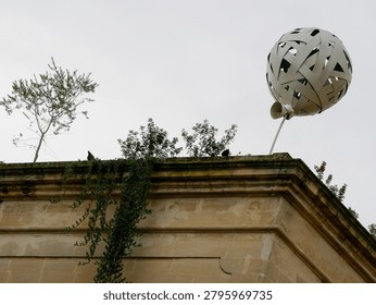 Low angle view of a unique modern balloon sculpture mounted on the roof of a historic stone building in Alberobello Italy featuring green rooftop plants climbing ivy and small trees