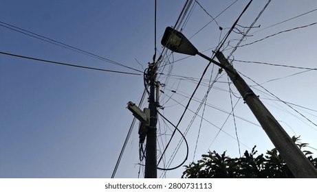Low angle view of utility pole with chaotic tangled electrical wires 
and street lamp against clear blue sky, with green tree visible in 
corner. Messy overhead power lines and cables on urban street pole 
in Southeast Asian city, concept of infrastructure challenges, 
urban development, and electricity distribution in developing countries.