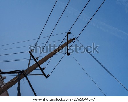 A low angle view of a utility pole and taut electrical wires diagonally crossing the frame against a deep blue sky, featuring a faint airplane contrail. It symbolizes connectivity, infrastructure, and travel.