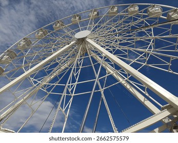 
Low angle view of a white Ferris wheel against a deep blue sky with scattered clouds, modern amusement ride under bright sunlight
